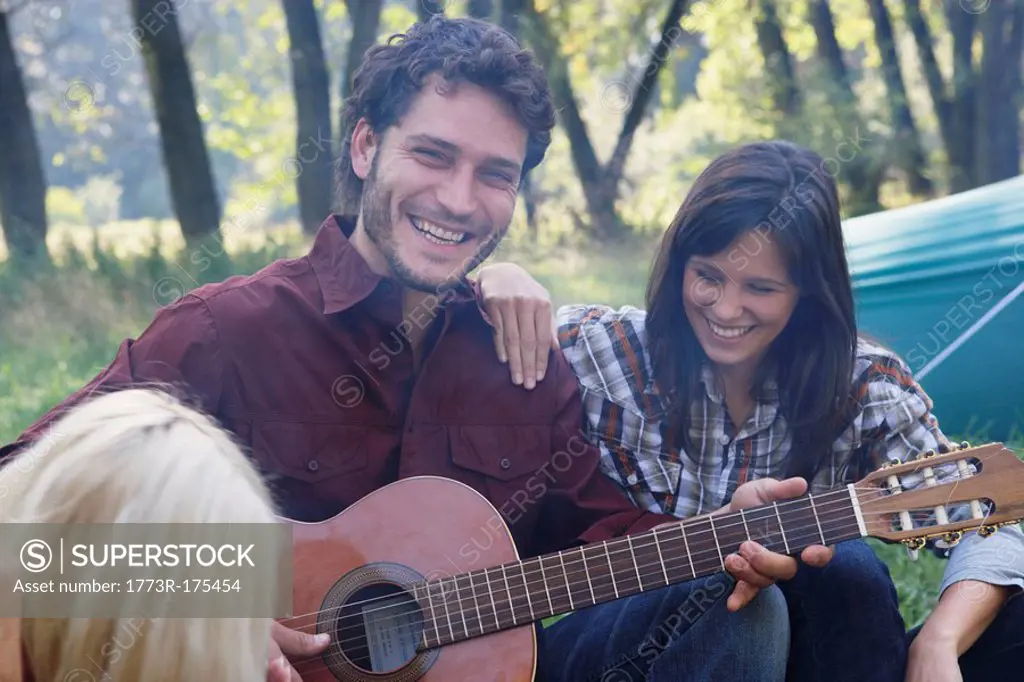 Man at campsite playing guitar with two woman listening and smiling
