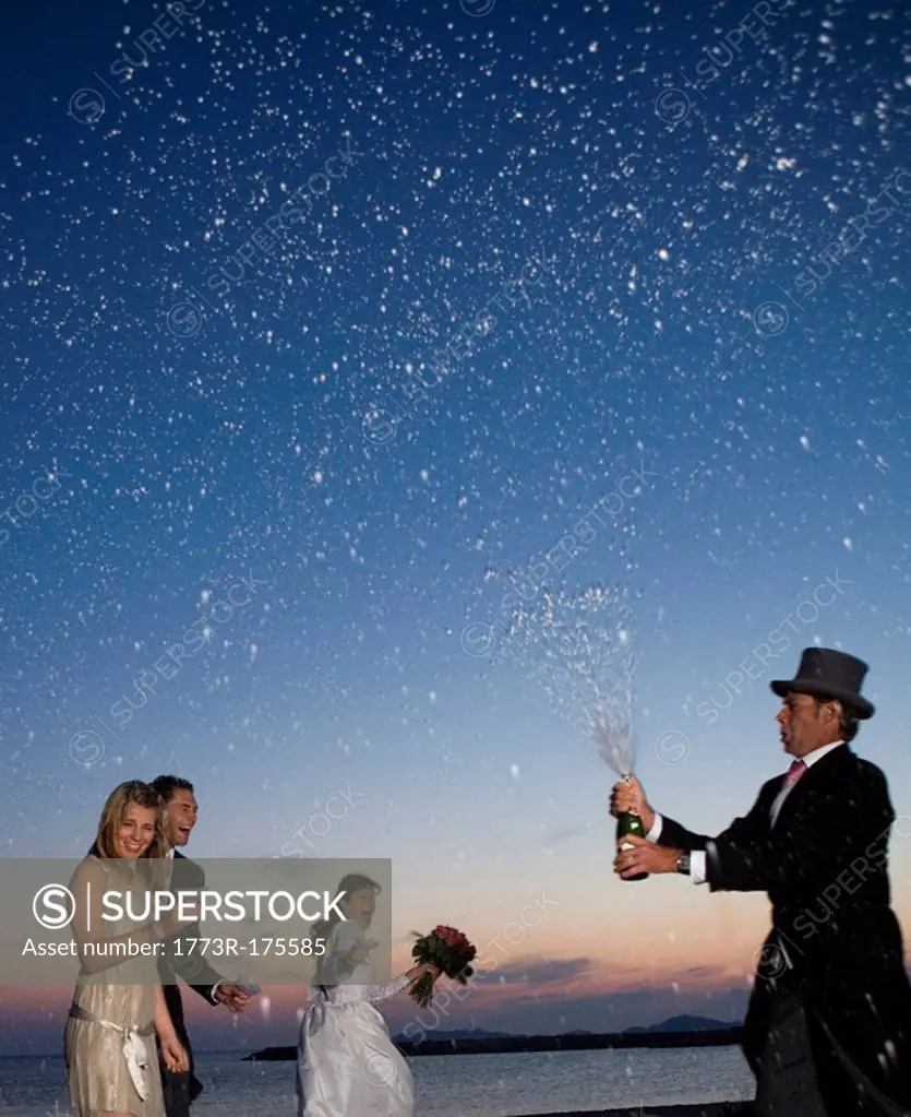 Man opening spraying bottle of champagne over wedding party on beach, sunset