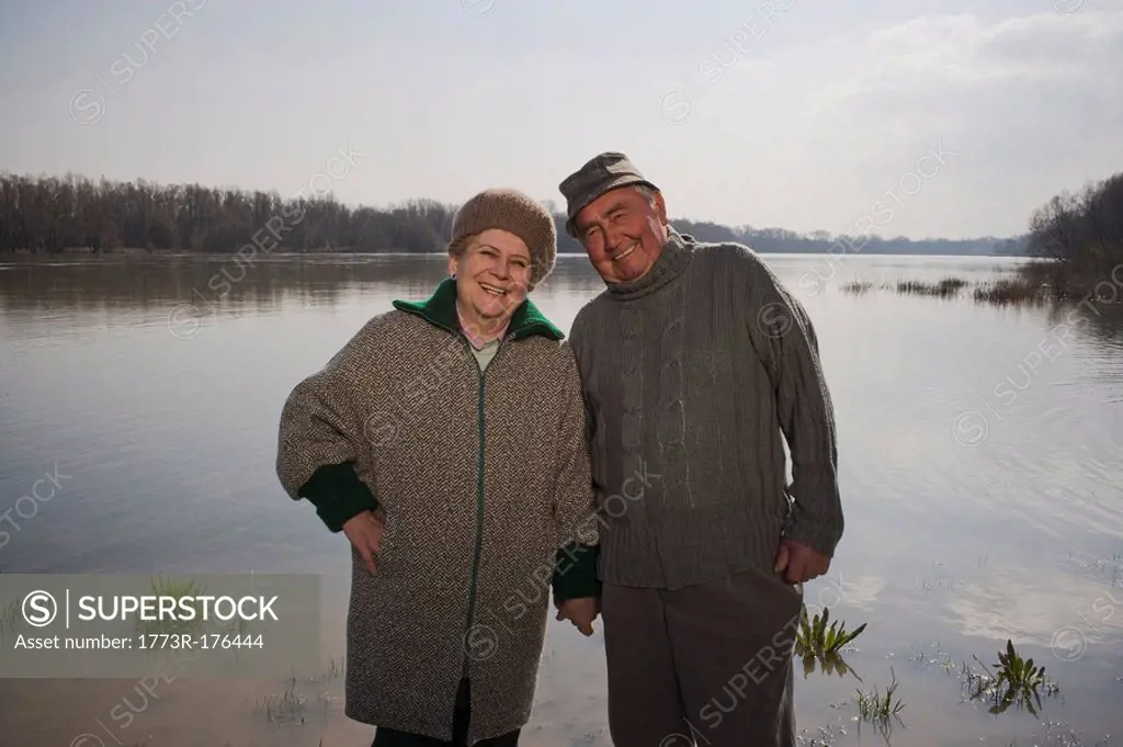 Senior couple standing by river, holding hands, smiling, portrait
