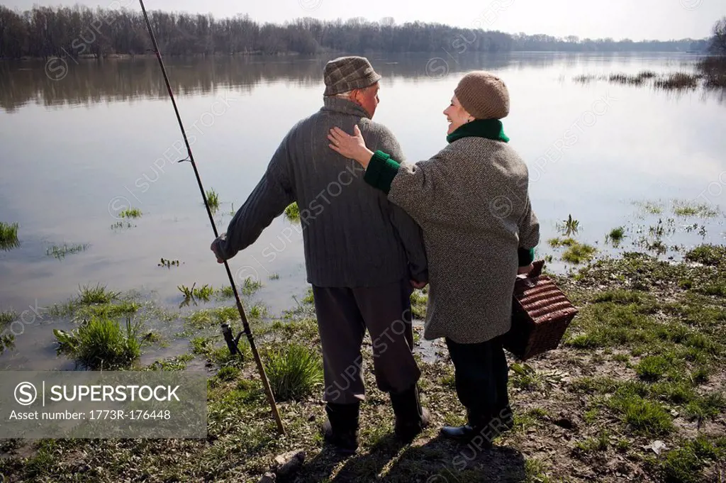 Senior couple standing by river, holding fishing rod and basket