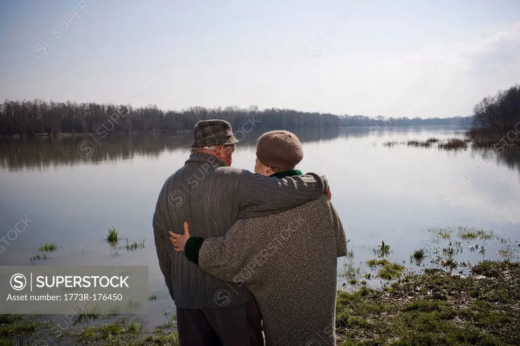 Senior couple standing by river, arms around each other, rear view