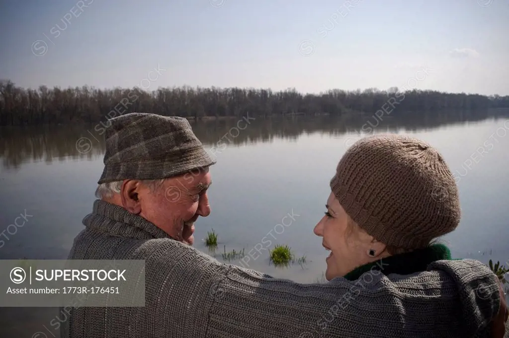 Senior couple by river, man holding arm around woman, rear view