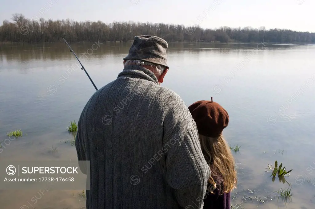 Grandfather teaching granddaughter 10-12 to fish in river, rear view