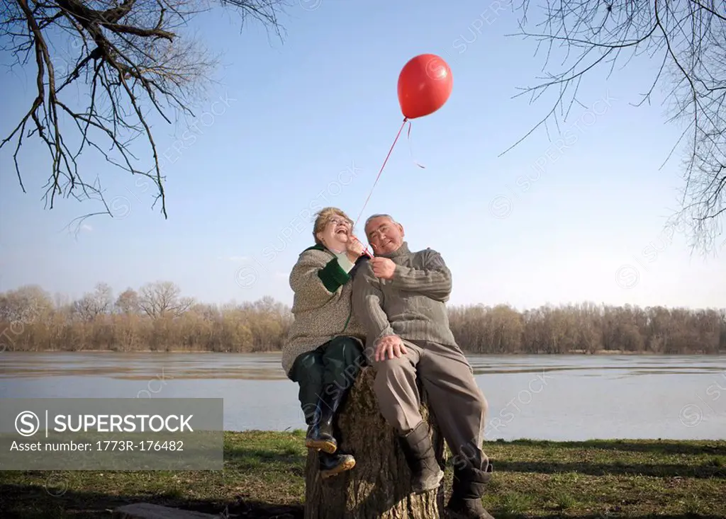 Senior couple sitting by river holding red balloon, smiling
