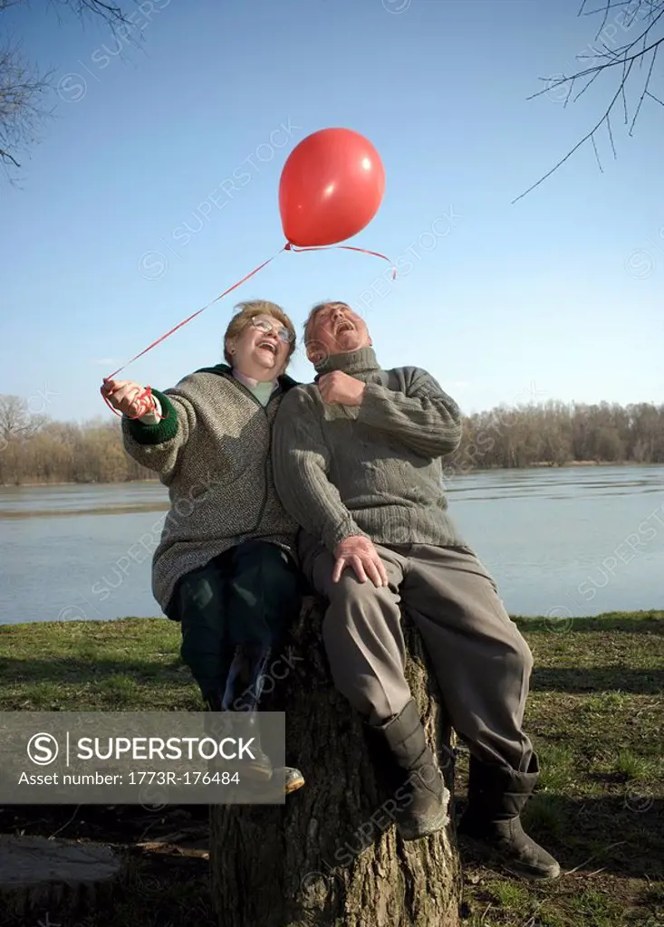 Senior couple sitting by river holding red balloon, smiling