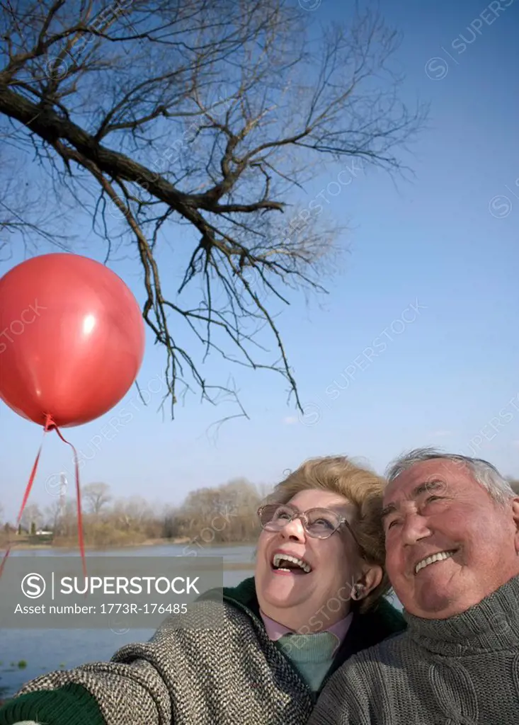 Senior couple by river holding red balloon, heads together, smiling