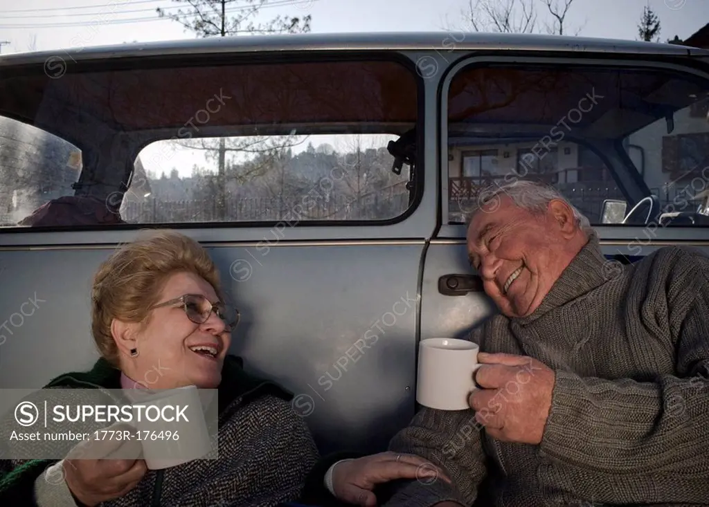Senior couple sitting by car in countryside, holding cups, smiling
