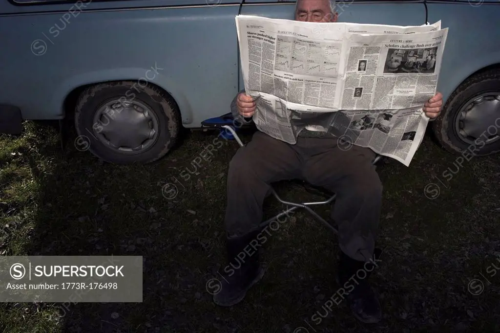 Senior man sitting by car reading newspaper