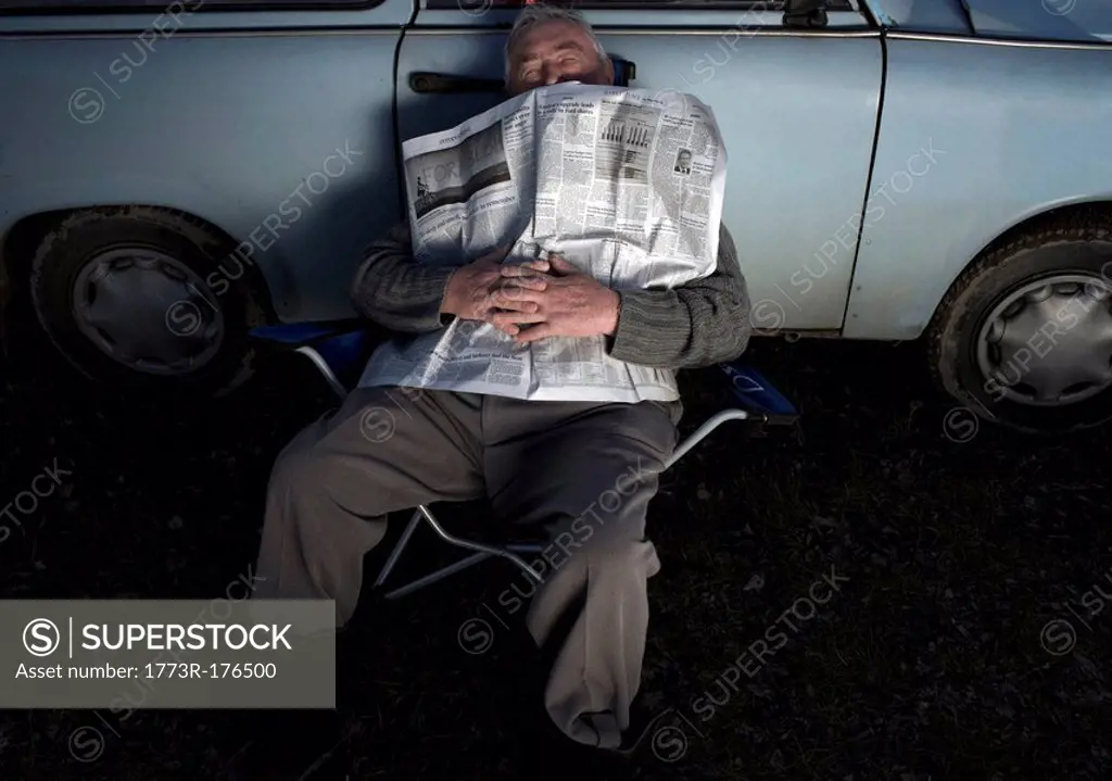 Senior man resting by car, holding newspaper, eyes closed