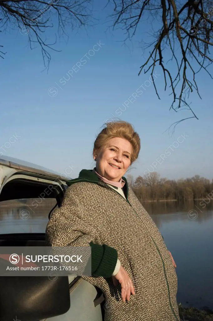 Senior woman leaning on car by riverside, smiling, portrait