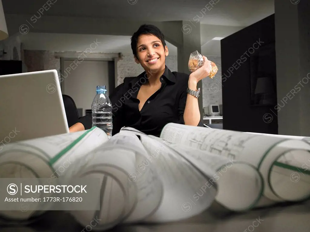 Female architect sitting at desk holding sandwich, smiling