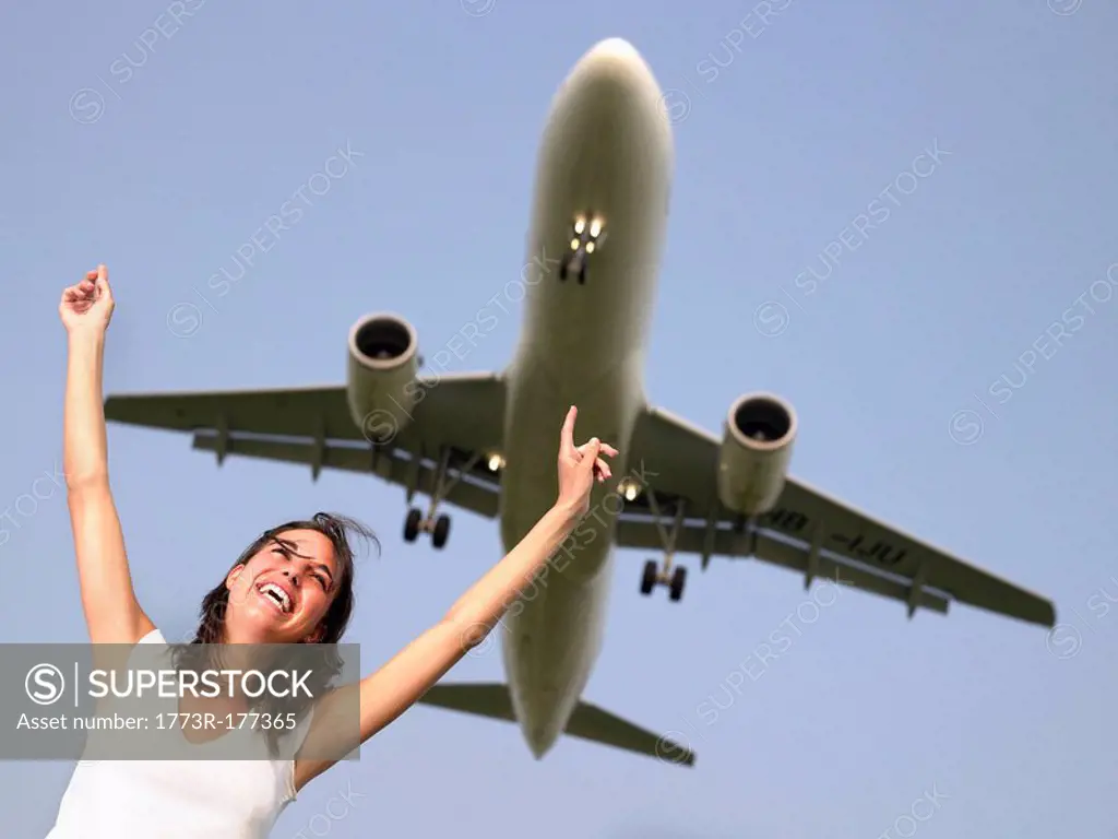 Woman smiling with arms outstretched standing below a plane flying overhead
