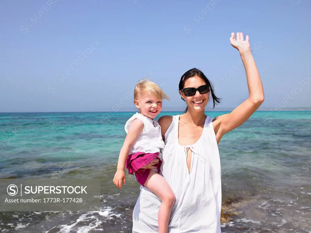Woman holding a young girl at the beach waving and smiling