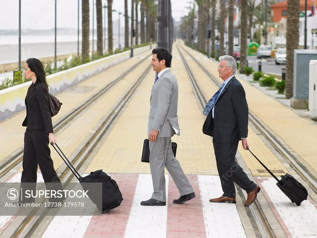 Businesswoman and two businessmen crossing double tram lines with suitcases at zebra crossing Alicante, Spain