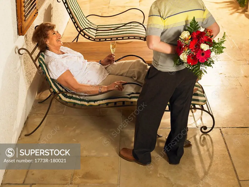 Senior woman lying on sun lounger smiling at senior man with flowers behind back