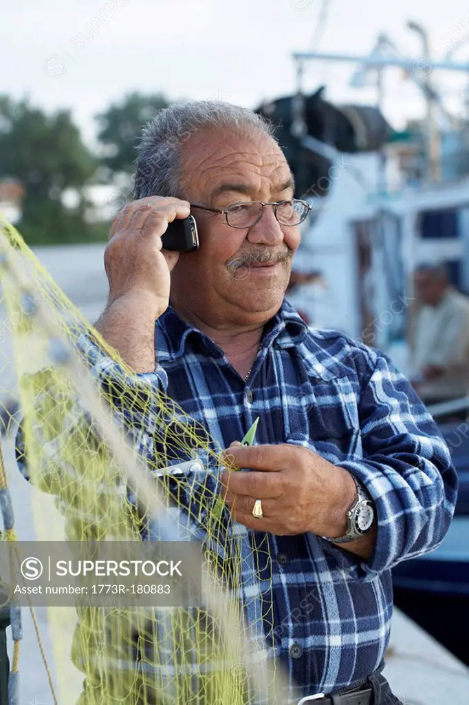 Mature fisherman fixing net while on cell phone
