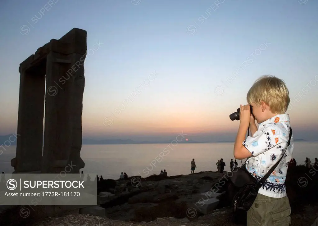 Young boy using binoculars to look at The Portara in Greece with tourists in background