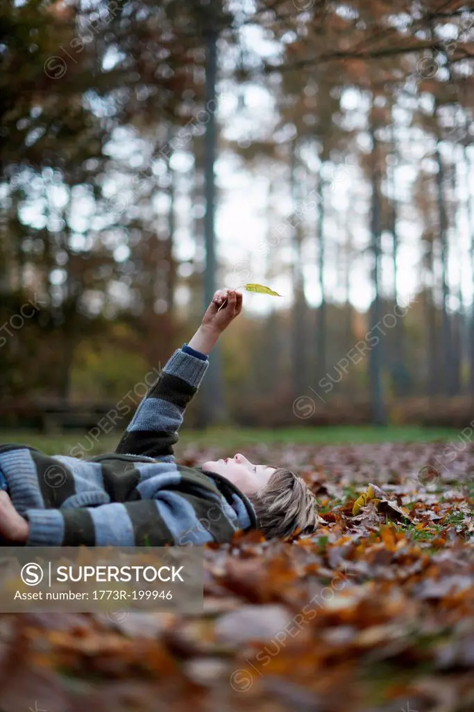 Boy lying on forest floor holding leaf in air