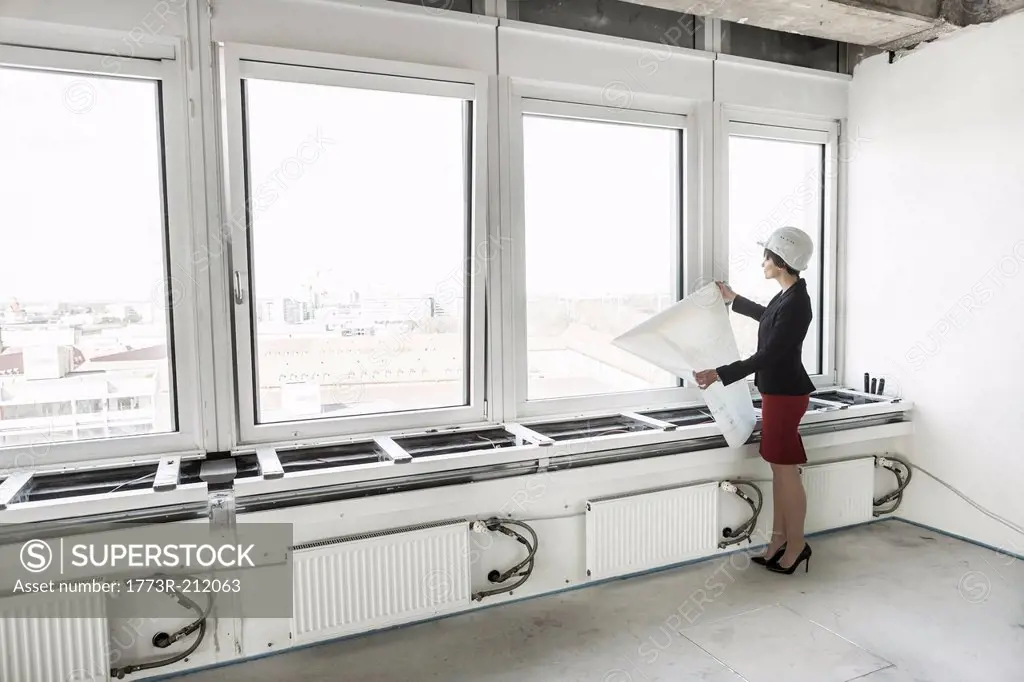 Businesswoman standing in empty unfinished office wearing hardhat and looking at plans