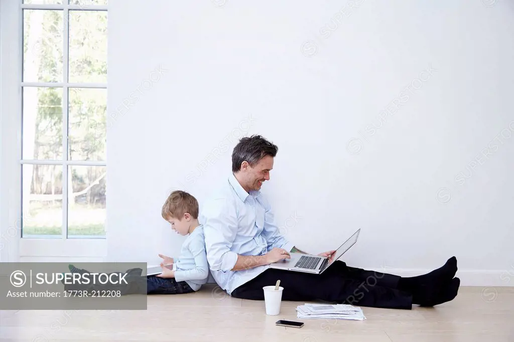 Father and son sitting back to back using laptop and digital tablet
