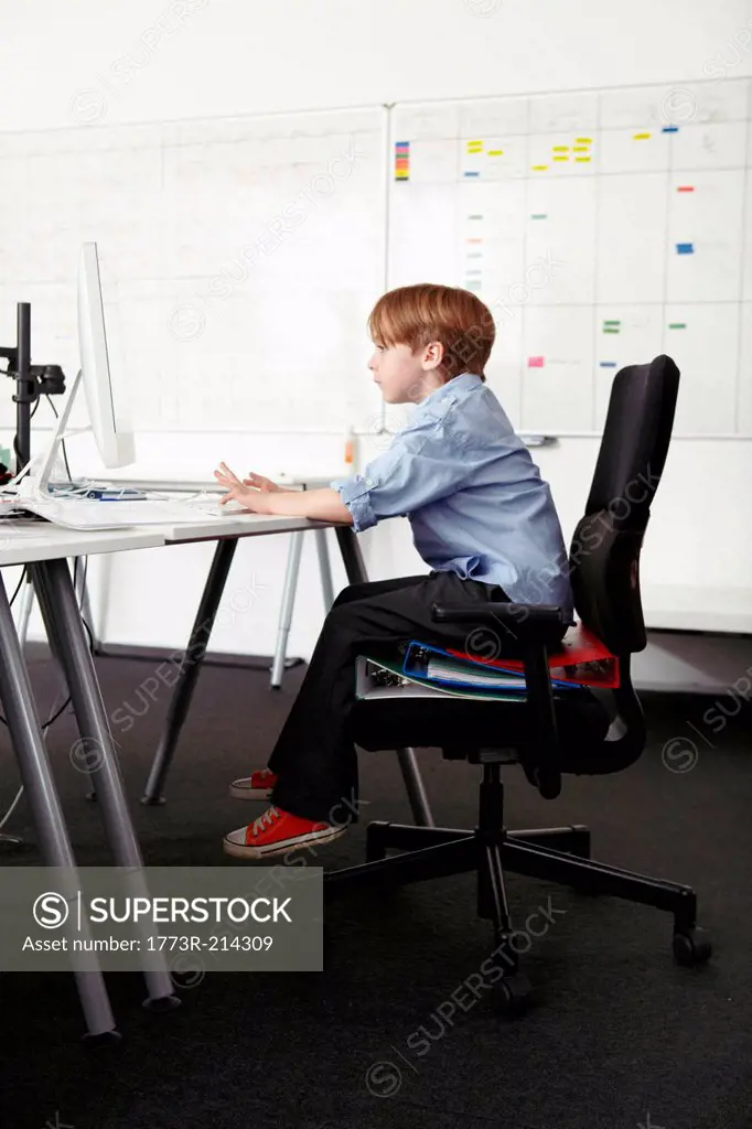 Boy sitting on ring binders on office chair using computer