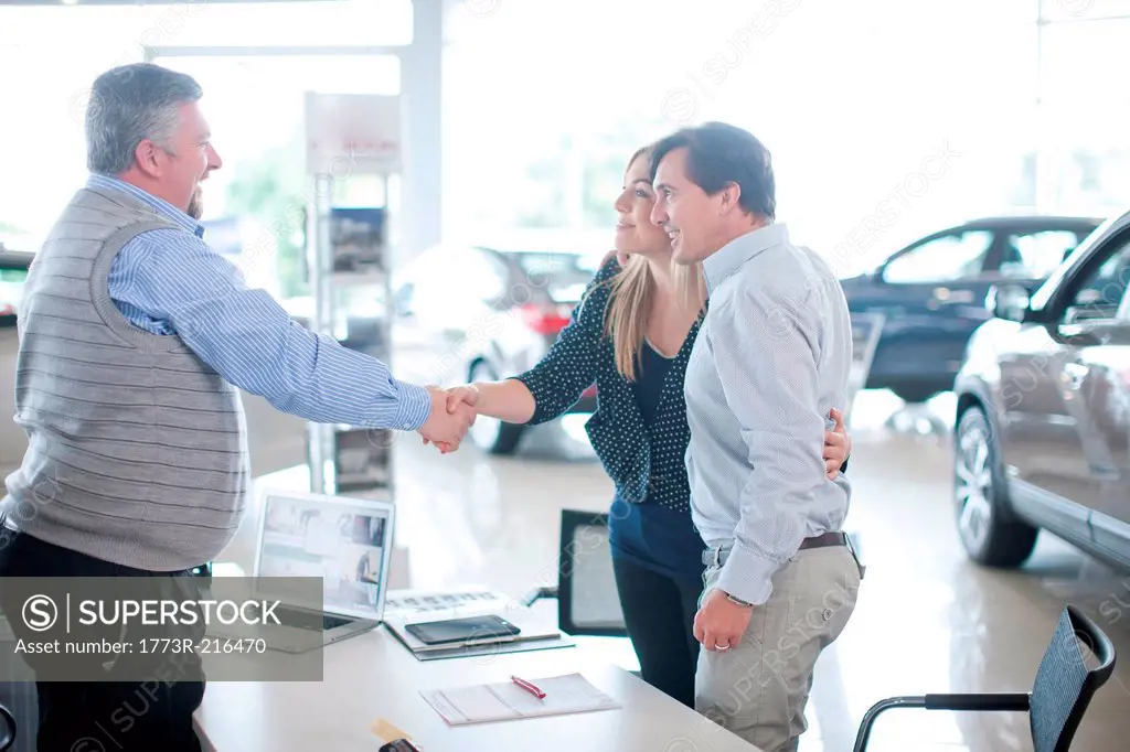Car salesman and couple shaking hands in car showroom