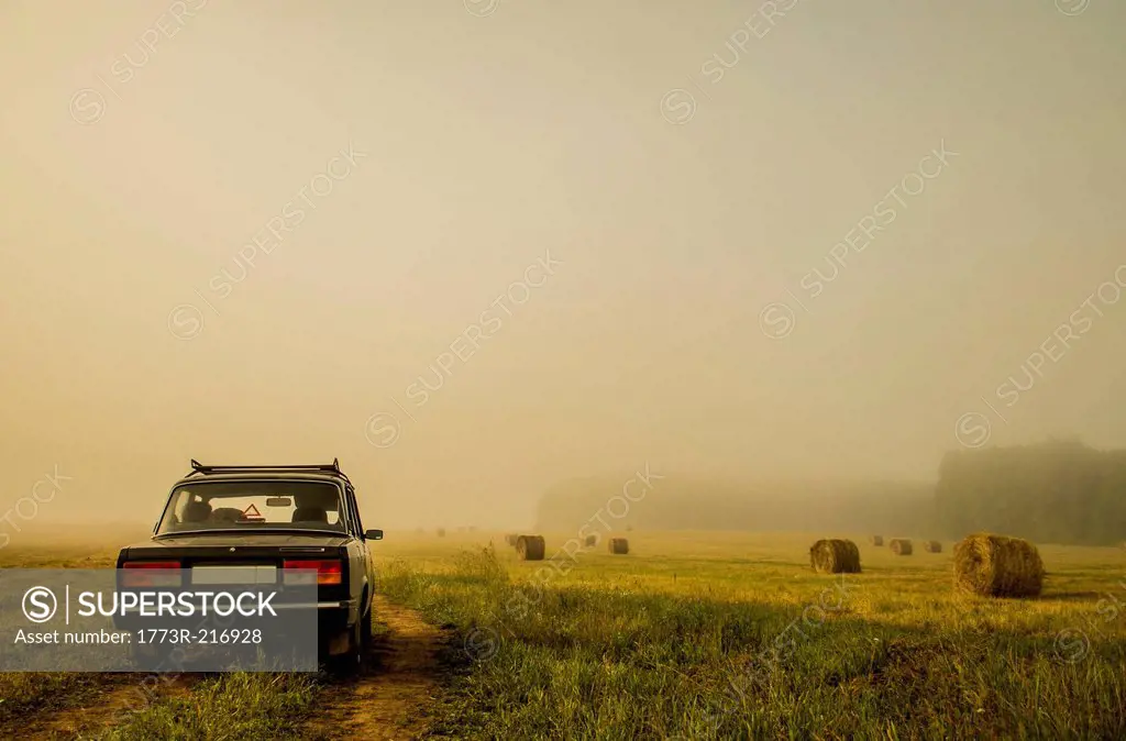 Car driving through field of haystacks on overcast day