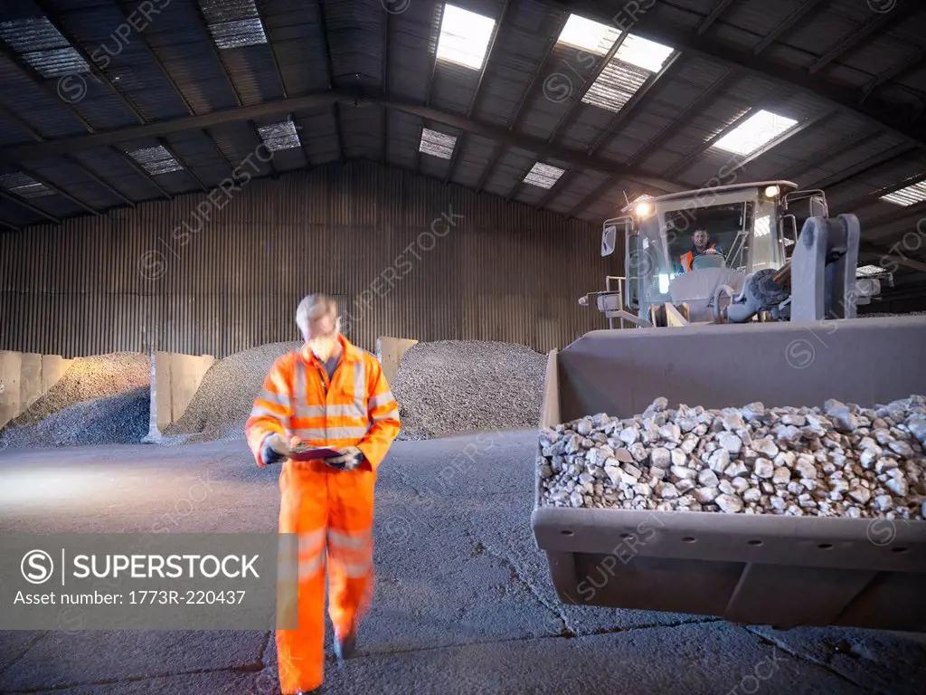 Worker checking clipboard in mineral store passing digger full of metal alloy