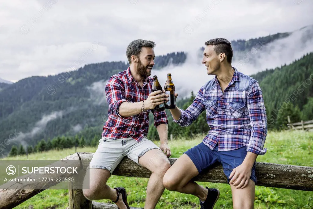 Two male friends drinking beer on fence, Tyrol Austria