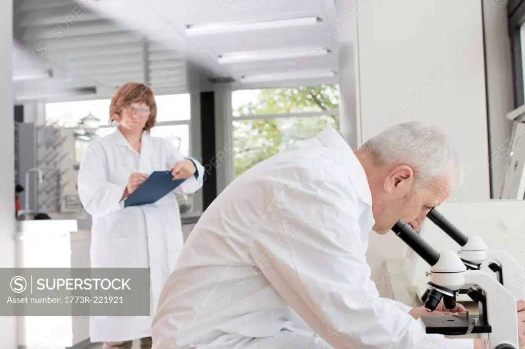 Scientists working in laboratory, man looking through microscope and woman taking notes
