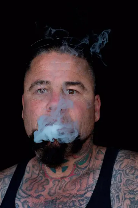 Portrait of bearded tattooed mature man looking at camera blowing smoke