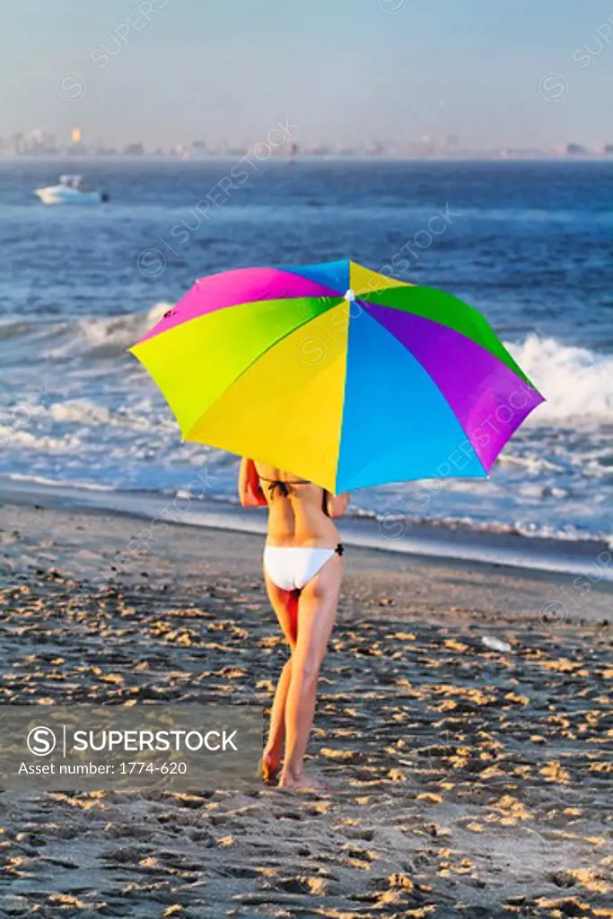 USA, New Jersey, Woman in bikini with beach umbrella standing on sandy Hook Beach and looking towards Staten Island and Manhattan