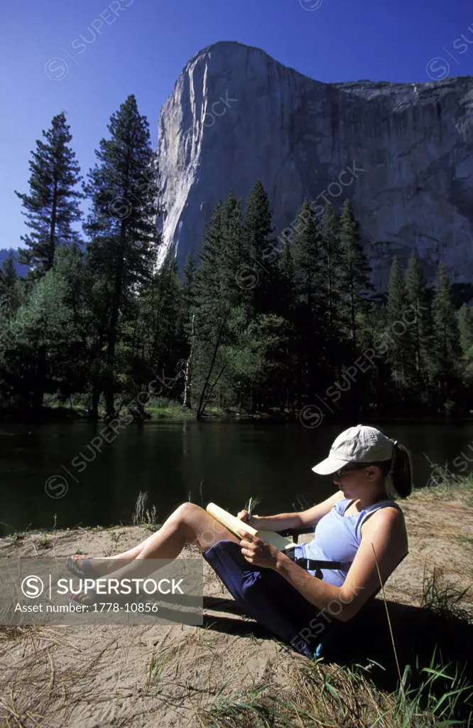 A woman writes in her journal while she sits next to a river in a valley in Yosemite National Park