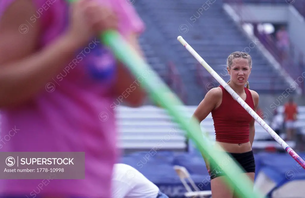 A teenage girl rests her pole on her shoulder in between pole vaulting jumps at a track meet