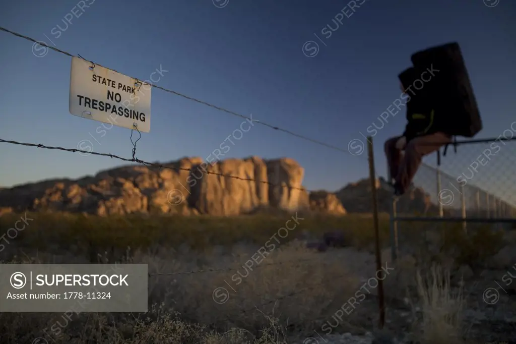 Man wearing a bouldering pad sits on a fence beside a sign