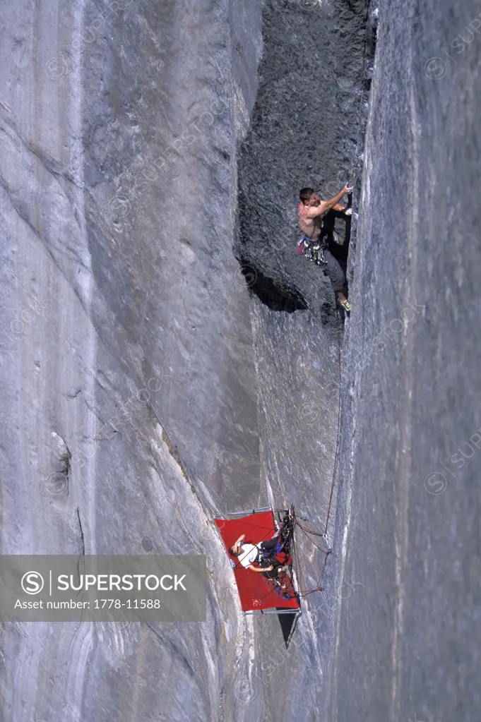 A woman belays a man rock climbing from a porta ledge on a big wall