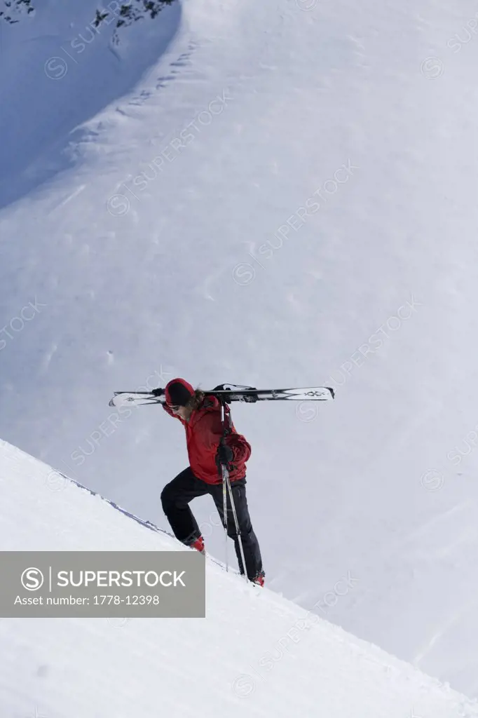 Skier carries skis up steep mountain on sunny blue sky day in Alaska backcountry