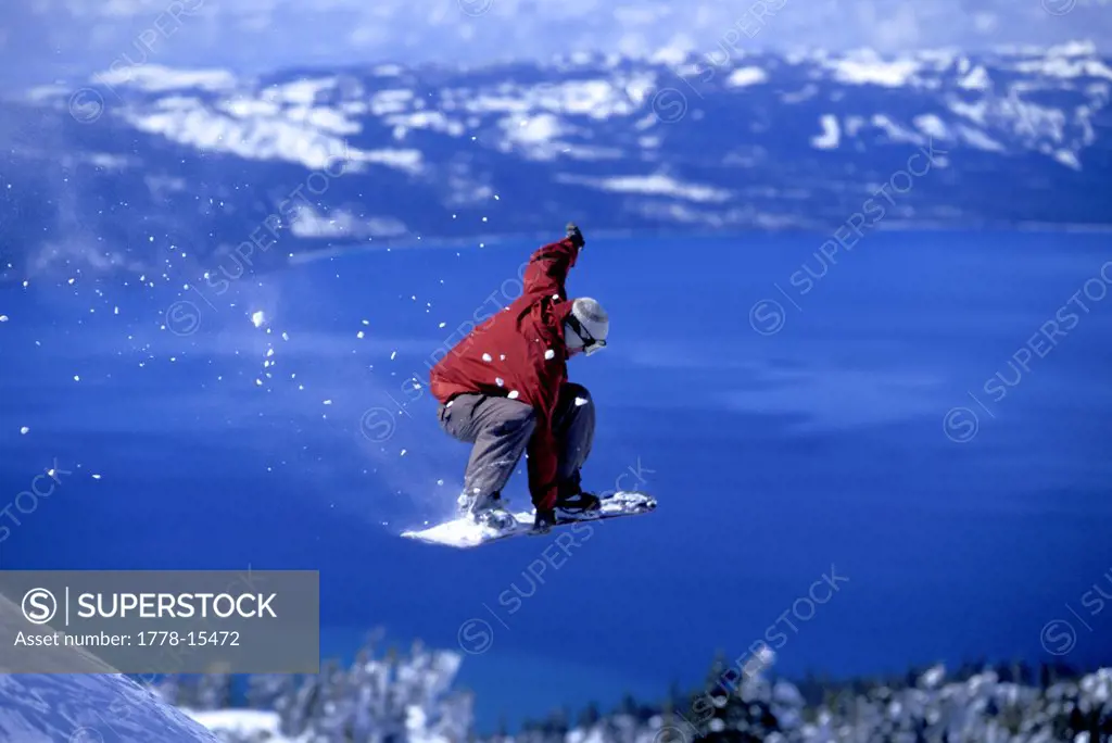 Snowboarder grabbing his board after jumping off of a cliff above a blue lake