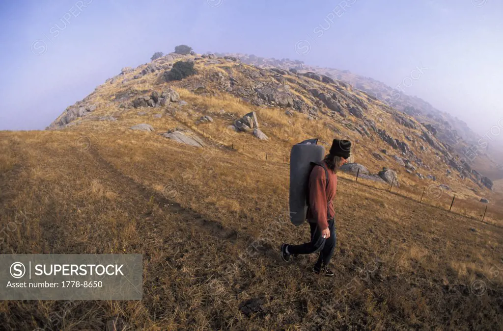 Man hiking to boulders with bouldering - rock climbing crash pad on his back  Exeter, California