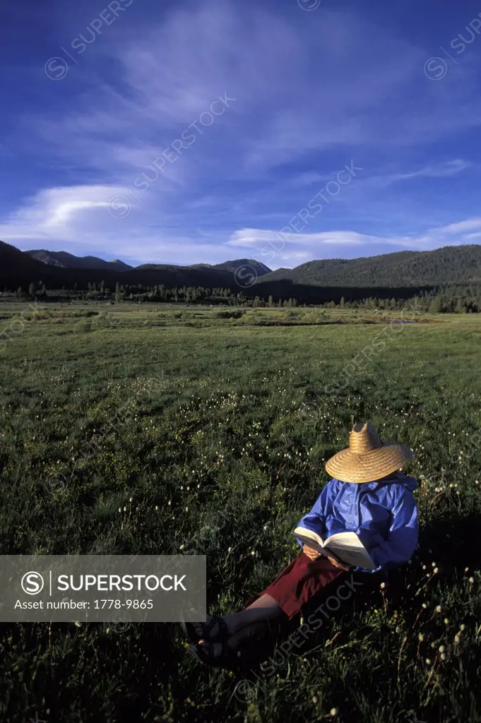 A woman reads a book in a straw hat while camping in a valley