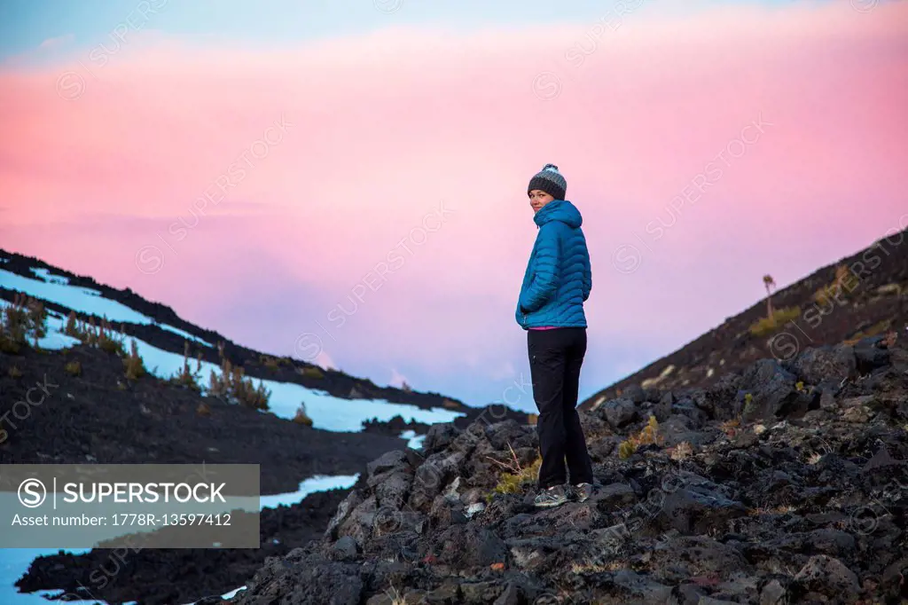 A woman in a teal parka stands in a high mountain environment after sunset.