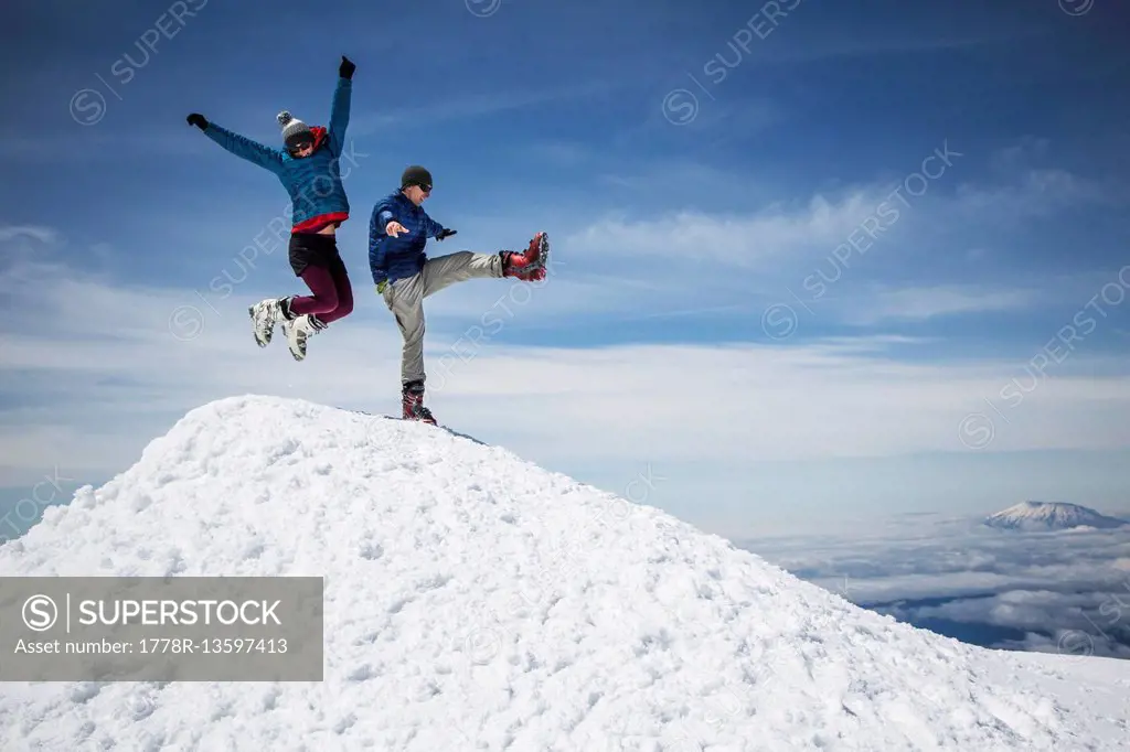 A woman jumps into the air and a man kicks one leg up in celebration of reaching a mountain summit.