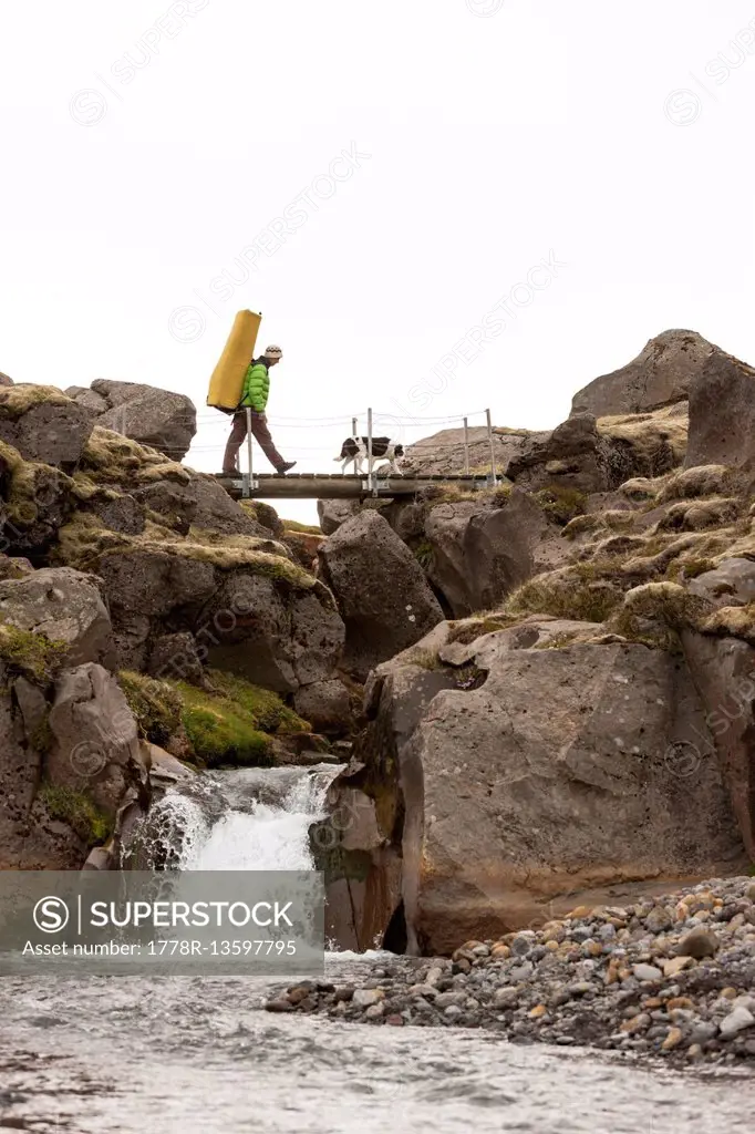 Climber with a bouldering matress on his shoulders walking over a bridge at Hnappavellir climbing area. Iceland.