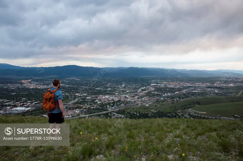 A man with a back pack looks over Missoula, Montana from the shoulder of Mount Jumbo at twilight.