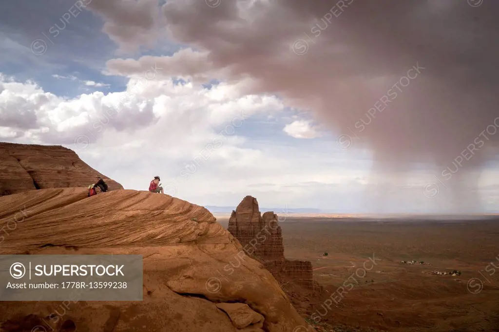 A woman sits on a cliff in the desert with a storm in the distance