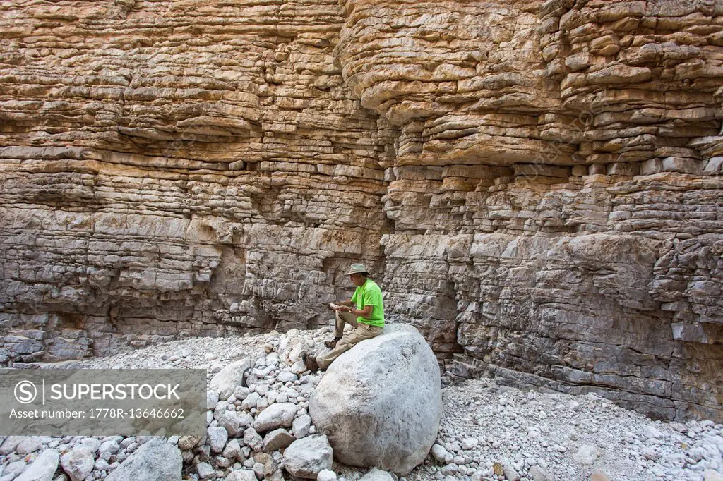 Senior Man Sitting On Rock In A Narrow Dry Canyon Of Steep Layered Rock Walls