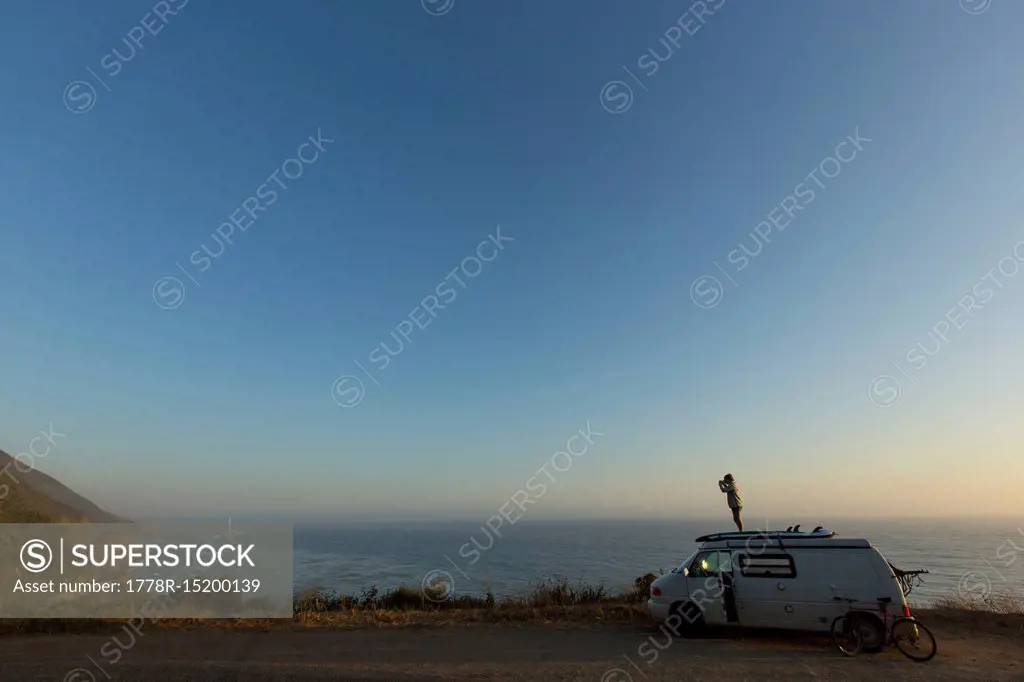 Girl standing on top of camper van while on road trip on Pacific Coast Highway and looking at view of Pacific Ocean, Big Sur, California, USA