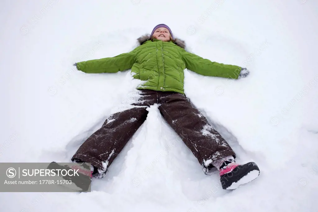 Young girl wearing a green winter jacket makes a snow angel in the snow.