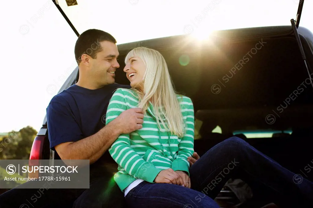 Couple enjoy themselves while sitting on the back of a truck in a parking lot.