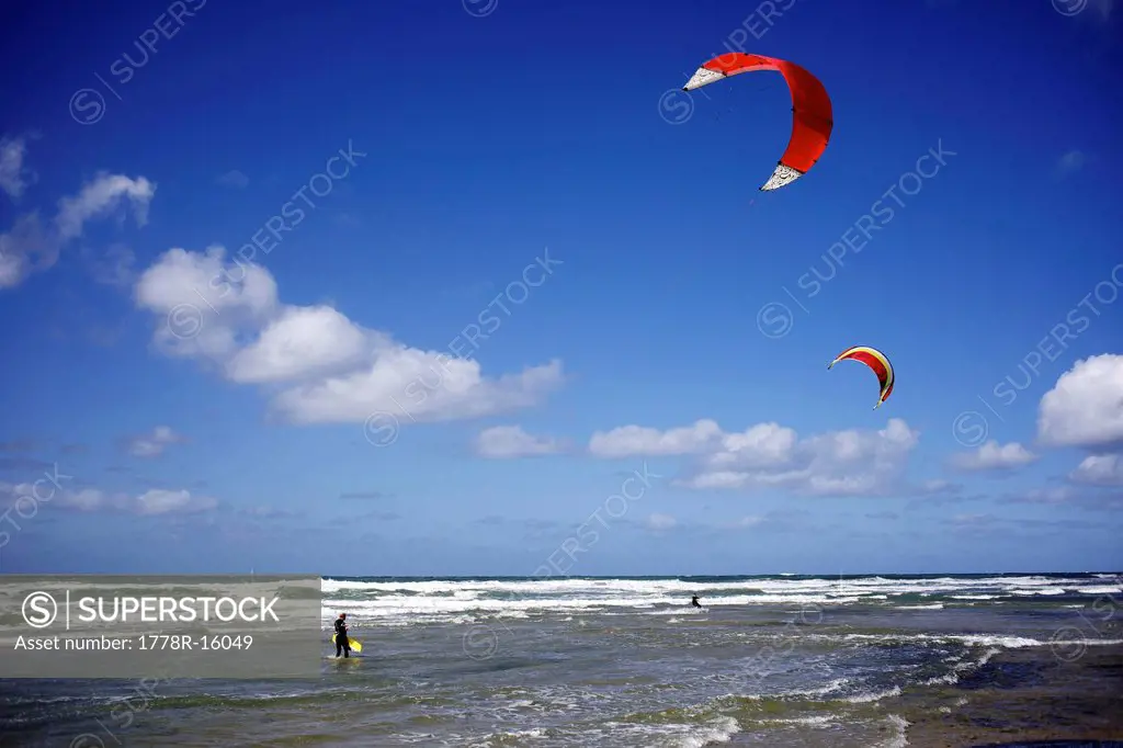 One male kitesurfer coming out of the water while a second male kitesurfer rides in the background.